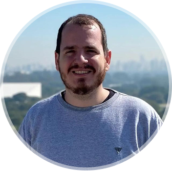 Foto de Rafael Faé sorrindo, usando moletom cinza, barba, com céu azul e árvores de fundo.
